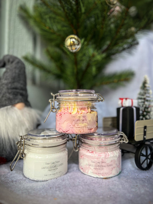 Three jars full of whipped soap sit in the foreground on a bed of snow. In the background is a Christmas wagon, a Tomte and a Christmas tree decorated with jingle bells in front of a grey wooden wall. 